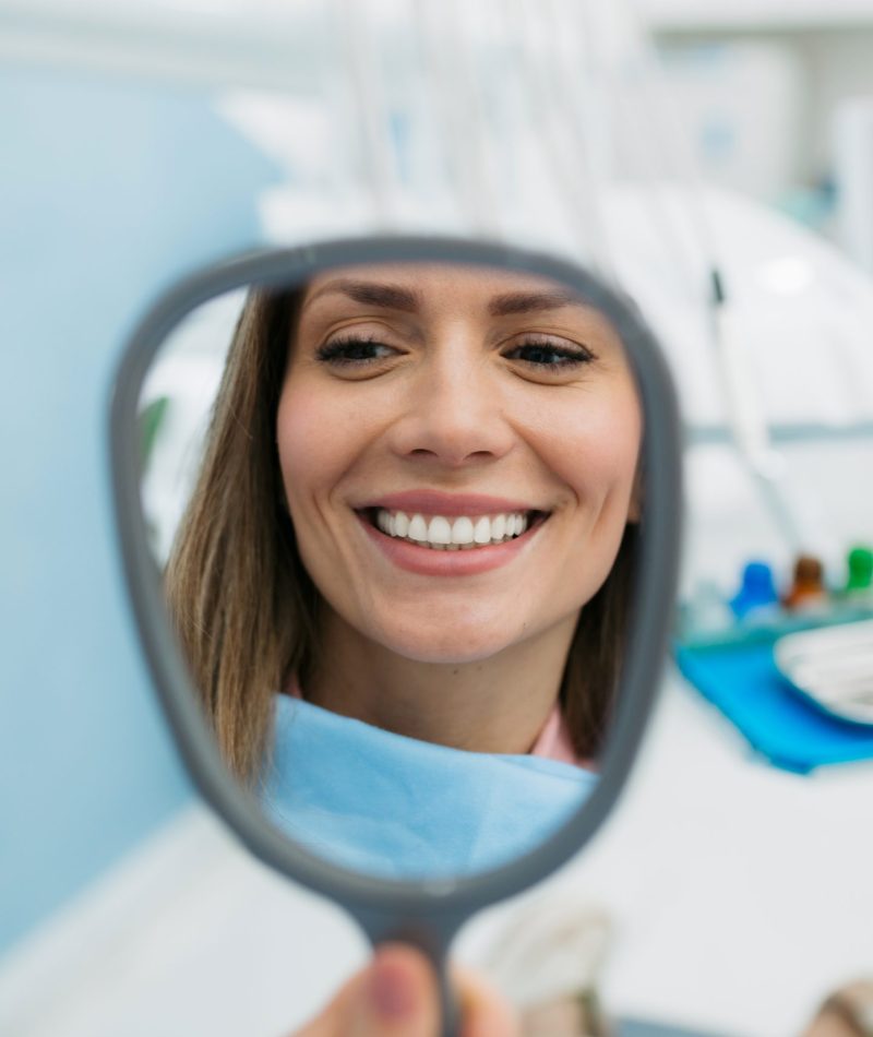 Young adult happy female patient relaxing while visiting a dentist office, holding a vanity mirror and with a toothy smile on her face inspecting her teeth